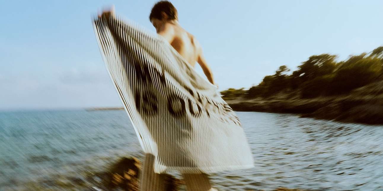 Adult male partially visible holding a large patterned beach towel near the ocean on a sunny day near a shoreline.