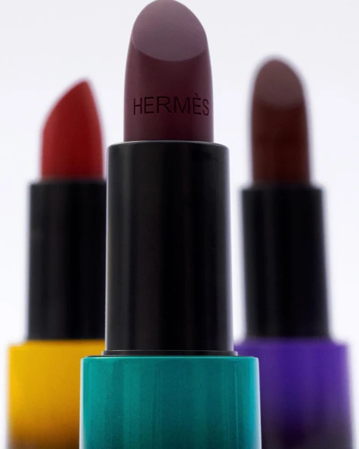 Close-up studio shot featuring three colorful lipsticks in focus, with Hermès branding visible on one. Bright, clean studio lighting.