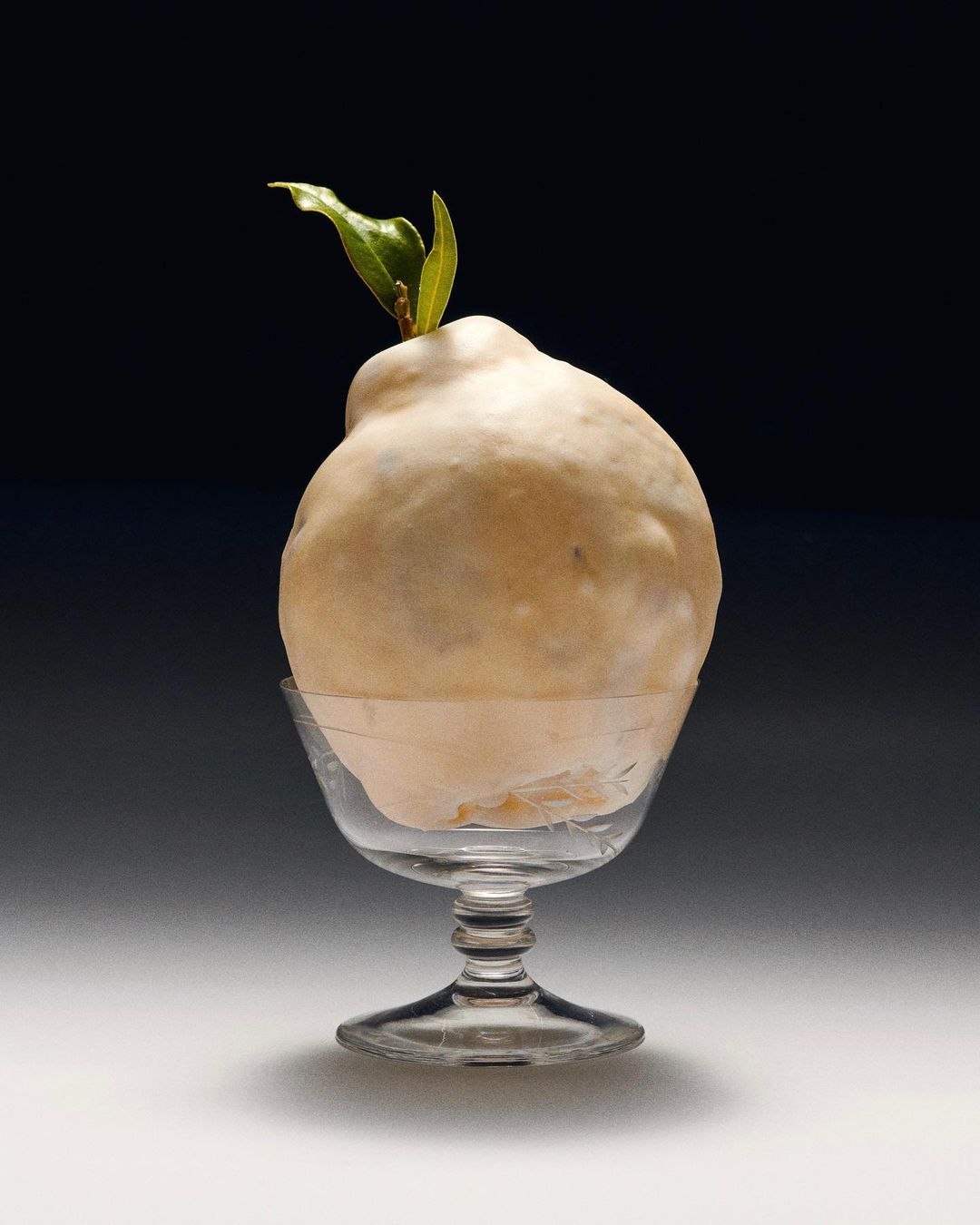 Quince fruit resting in a clear glass cup. The fruit has a small stem with green leaves, set against a gradient black background.