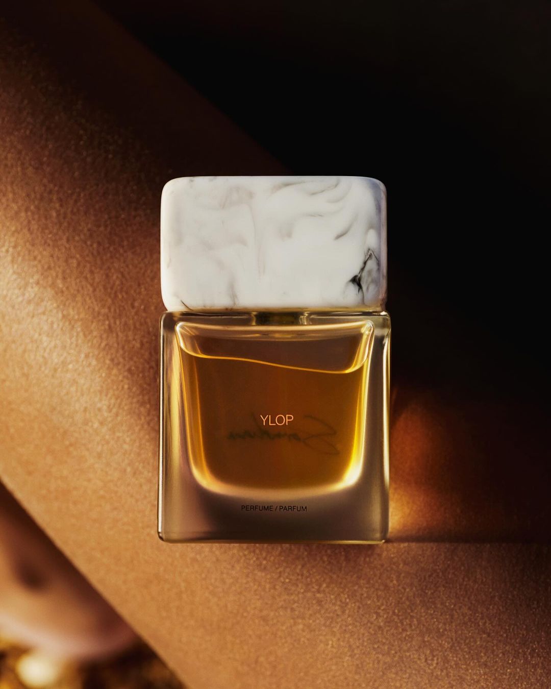 A rectangular perfume bottle with a white marble cap resting against skin in warm lighting
