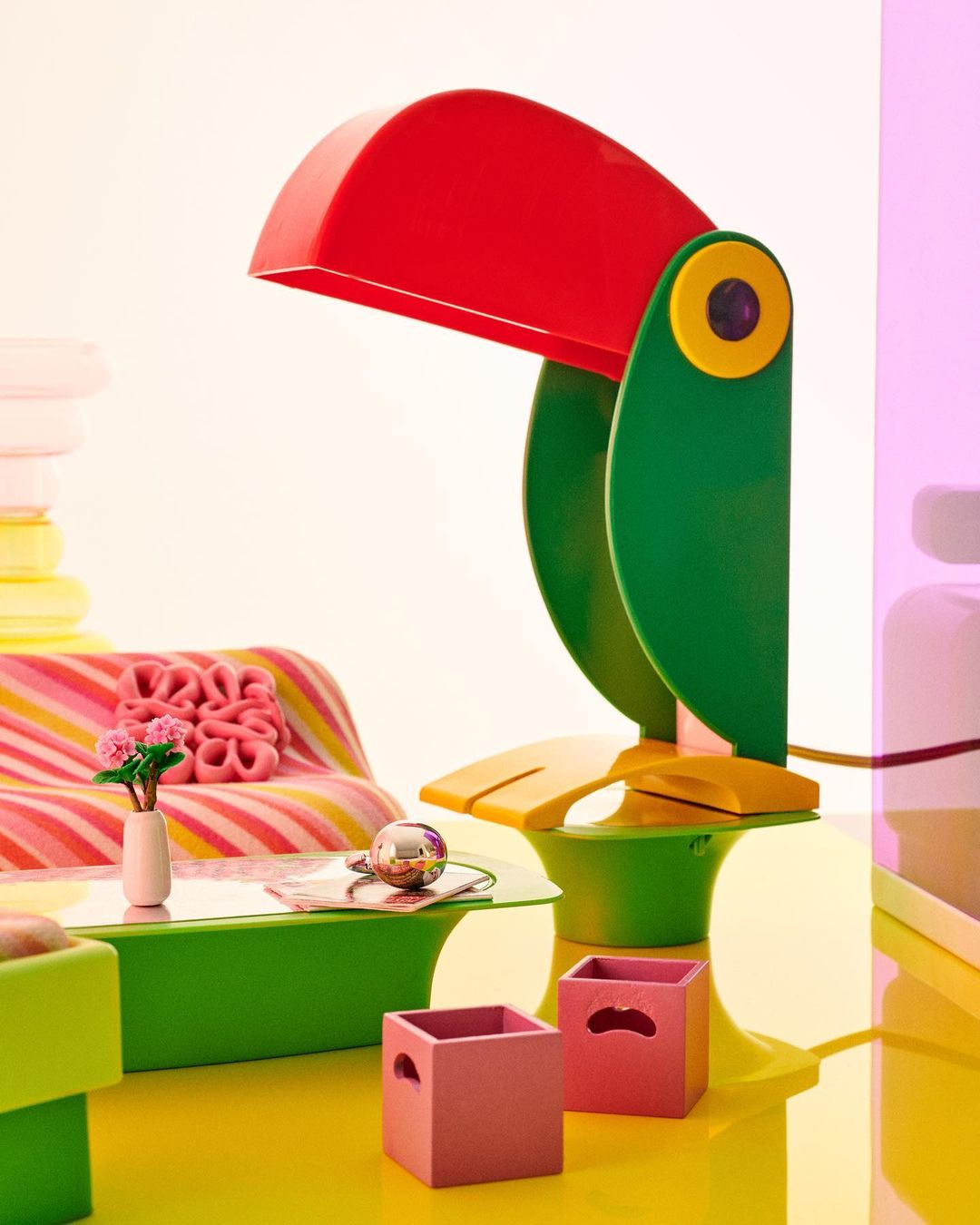 Stylized product shot features a colorful toucan lamp on a green table, surrounded by pink storage boxes, flowers, and vibrant hues.