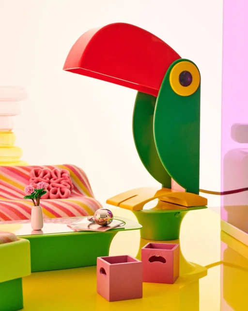 Stylized product shot features a colorful toucan lamp on a green table, surrounded by pink storage boxes, flowers, and vibrant hues.