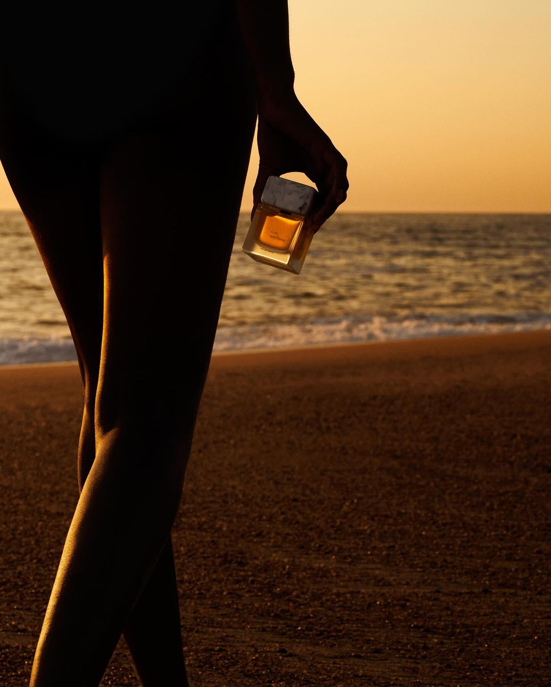 Silhouette of a person's legs and arm holding a perfume bottle on a beach at sunset with golden light, ocean waves, and sand.