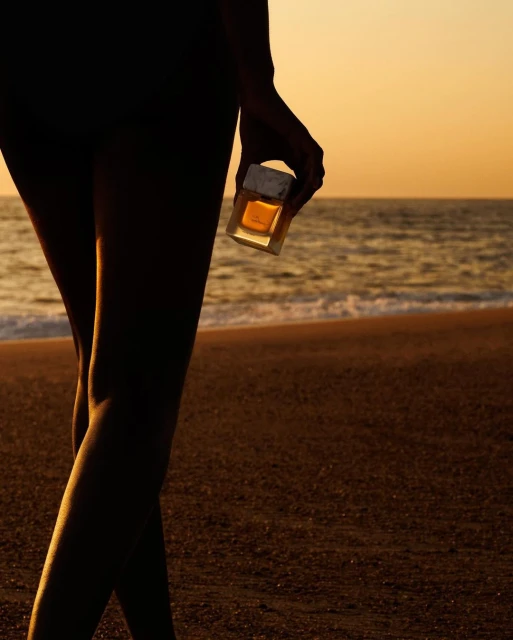 Silhouette of a person's legs and arm holding a perfume bottle on a beach at sunset with golden light, ocean waves, and sand.