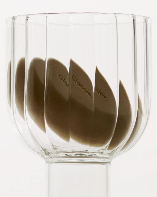 Cosmetic sponges with the Glossier logo in a clear ribbed glass bowl on a white background in a close-up shot. Beauty product photography.