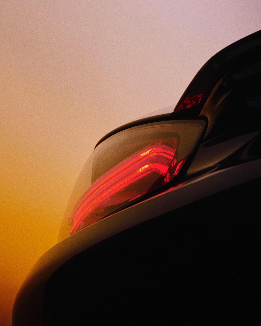 Close-up of a sleek black car's glowing red taillight against a warm sunset-toned sky creating a modern, automotive aesthetic.