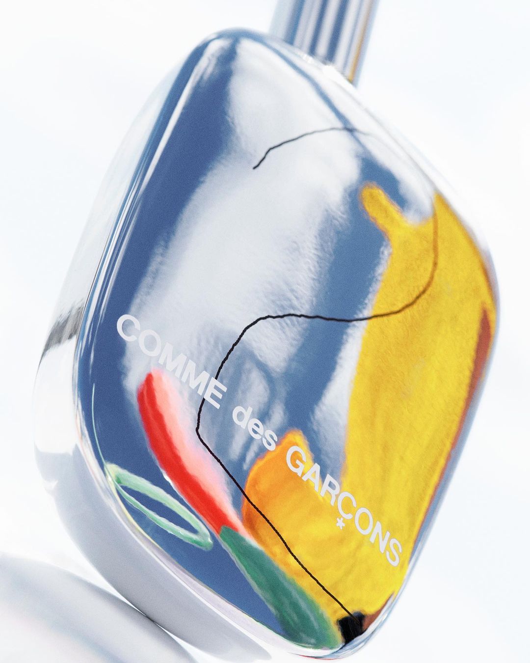 A glass perfume bottle by Comme des Garçons, featuring colorful abstract art and the brand name, set against a blurred white backdrop.