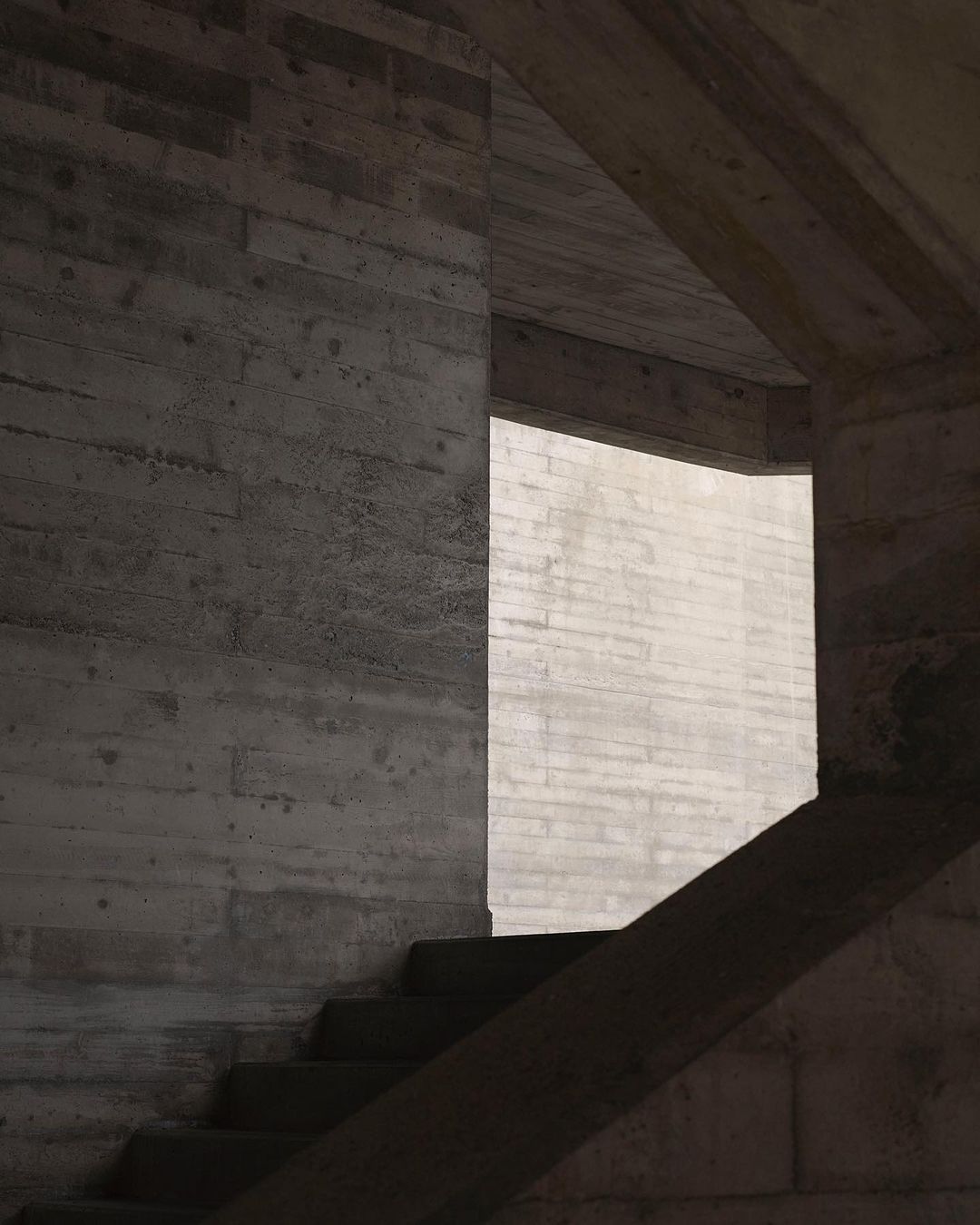 Architectural shot featuring concrete staircase and interior wall with natural light in a minimalist style; grey color palette.
