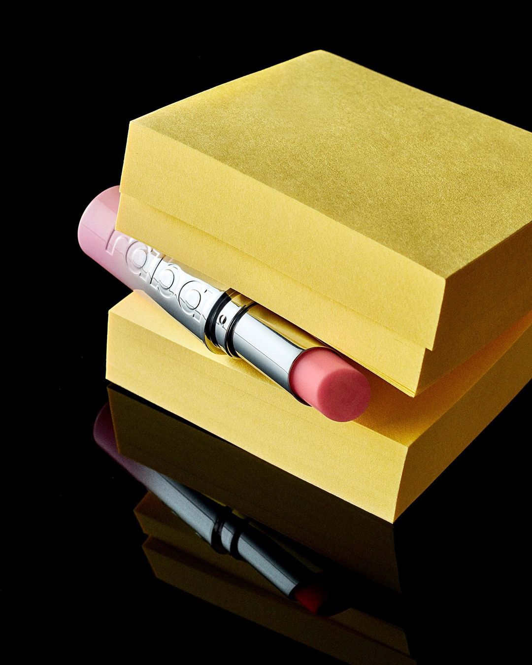 Close-up studio shot of pink lipstick tube on stacked yellow blocks against a black backdrop, with reflection showing on the surface.