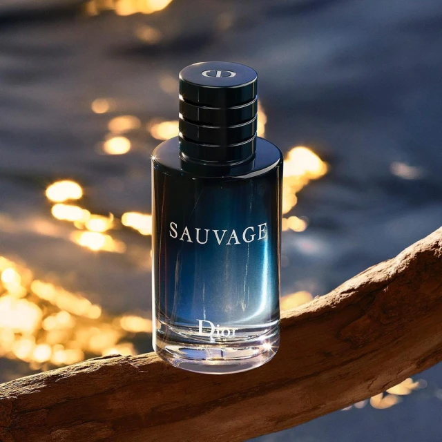Fragrance bottle of Dior Sauvage sits on a piece of driftwood with blurred background of water reflecting sunlight.