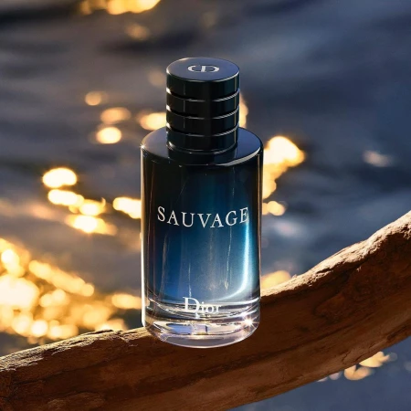 Fragrance bottle of Dior Sauvage sits on a piece of driftwood with blurred background of water reflecting sunlight.