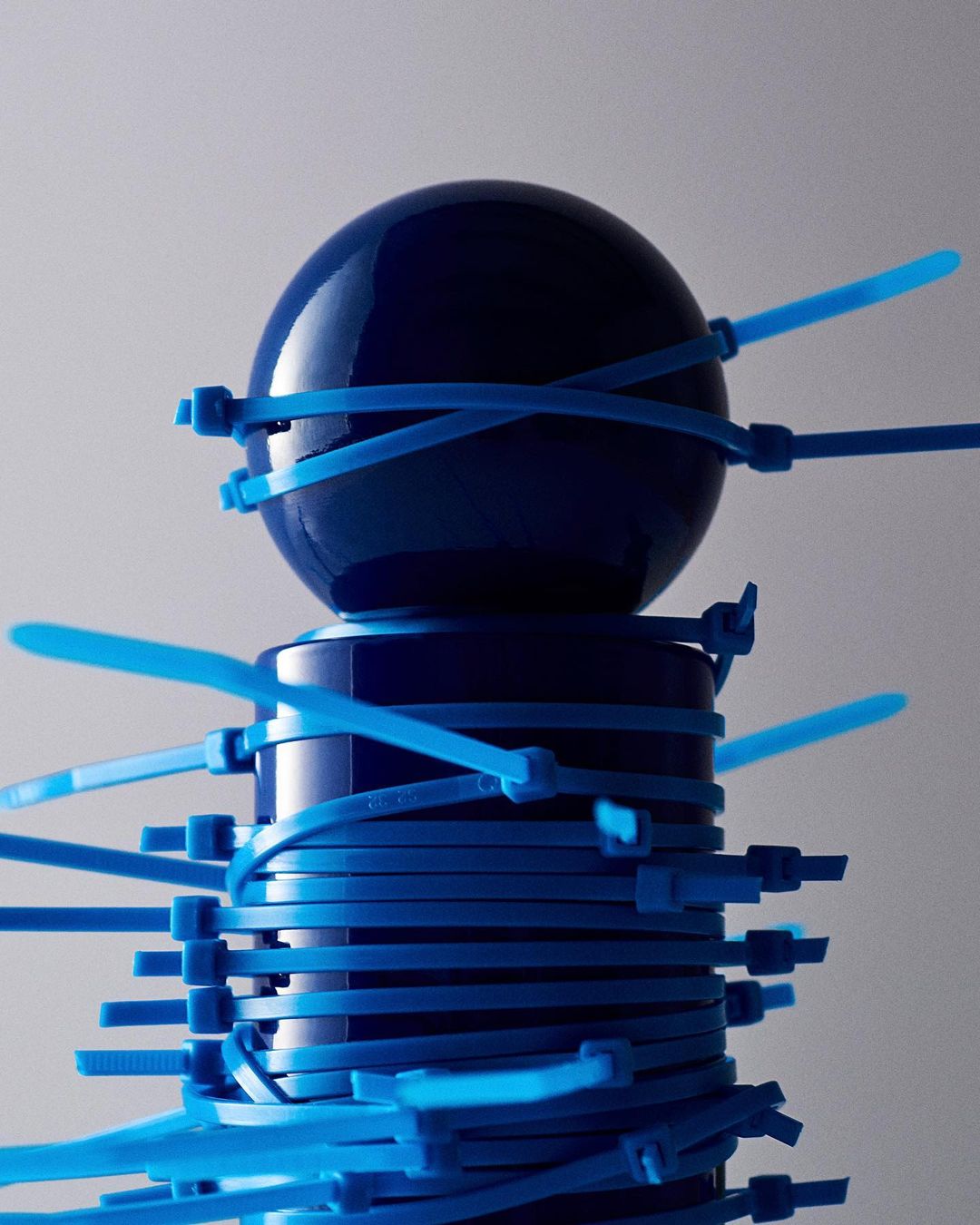 A blue ball sits atop a blue container, both secured by several light blue zip ties against a gray backdrop in a studio setting.