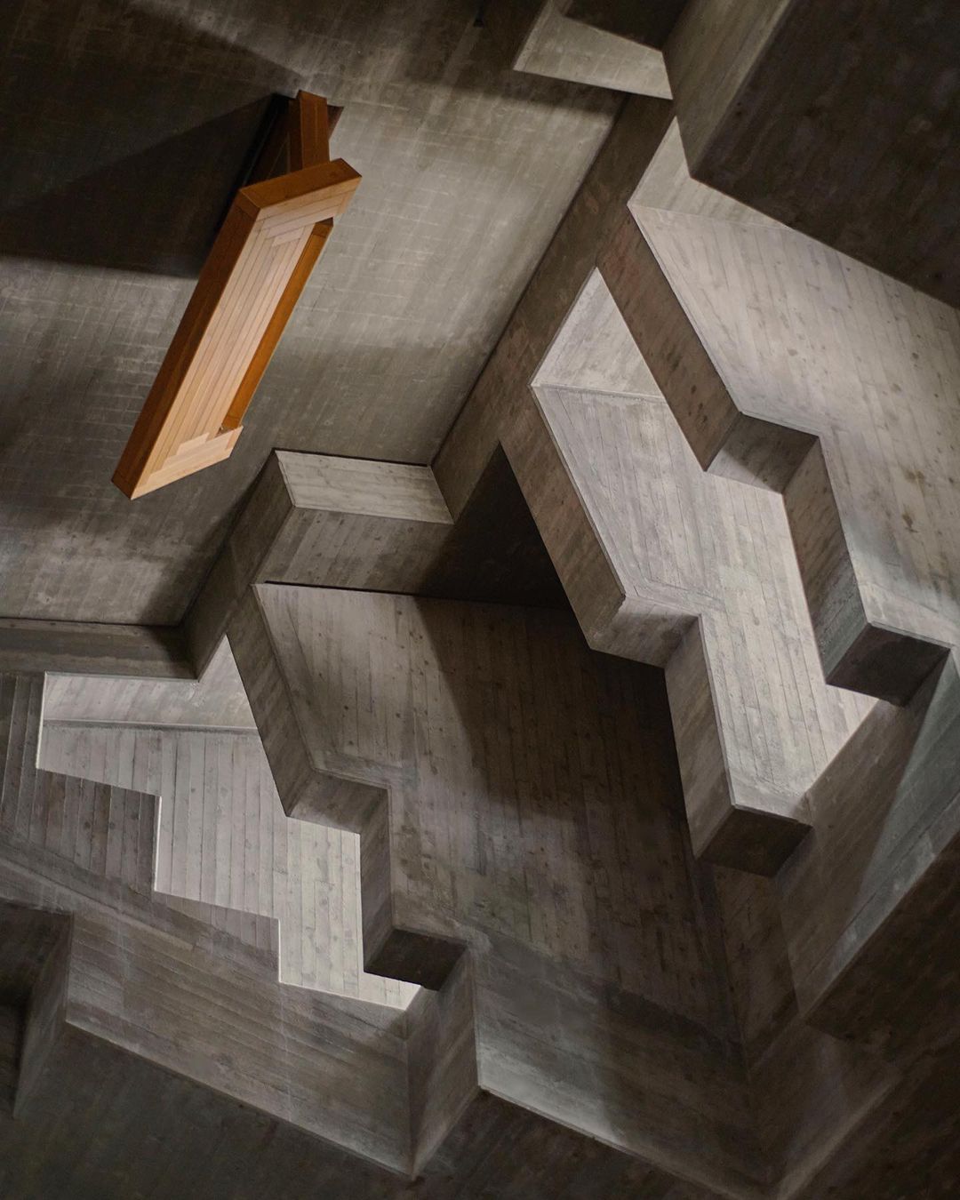 Concrete architectural ceiling with geometric patterns, featuring an overhead wooden light fixture in a neutral-toned, indoor space.