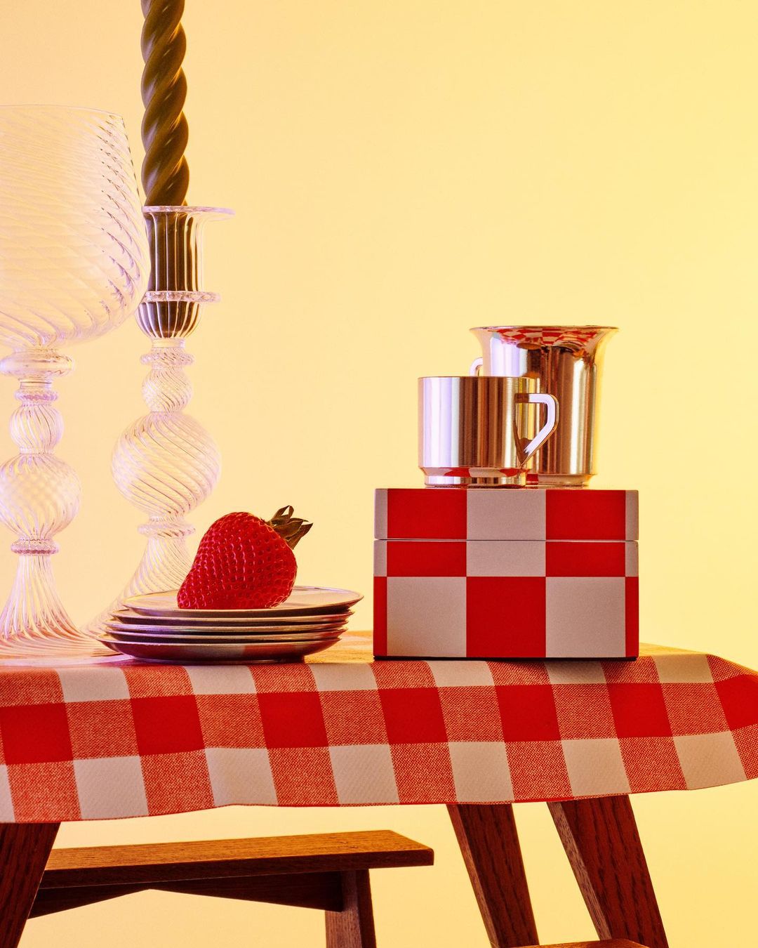 Still life of strawberry on plates, glassware, silver mugs, and a checkered box atop a red and white checkered tablecloth.