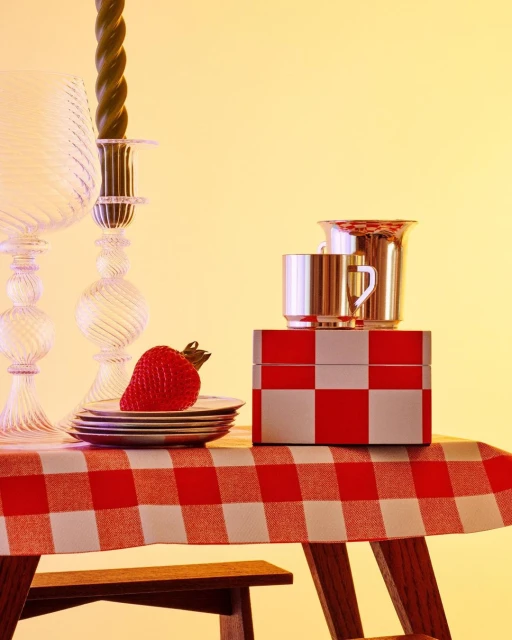 Still life of strawberry on plates, glassware, silver mugs, and a checkered box atop a red and white checkered tablecloth.