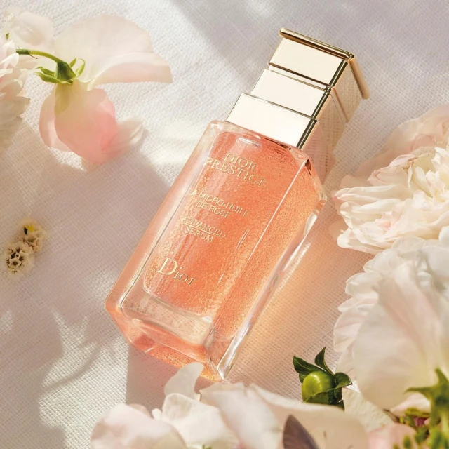 Dior Prestige serum bottle on white fabric surrounded by white flowers, showcasing luxury skincare and beauty product presentation.