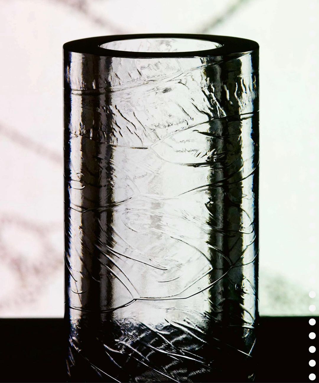 Cylindrical clear glass vase with textured design and glossy reflection on black surface against a blurred, bright background.