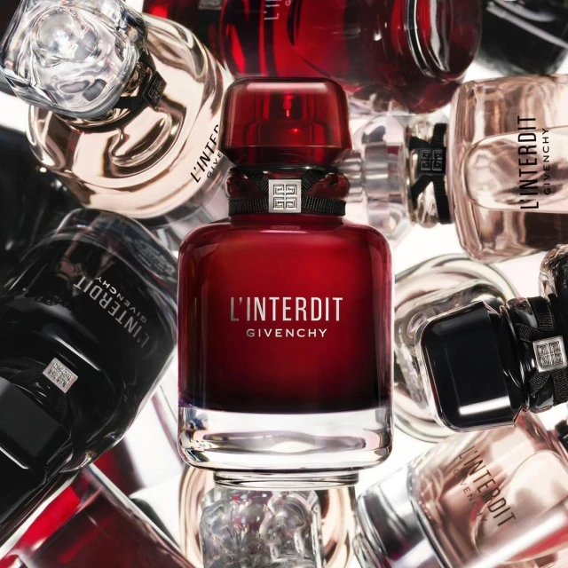 Close-up of a red Givenchy L'Interdit perfume bottle surrounded by other assorted fragrance bottles on a white background.