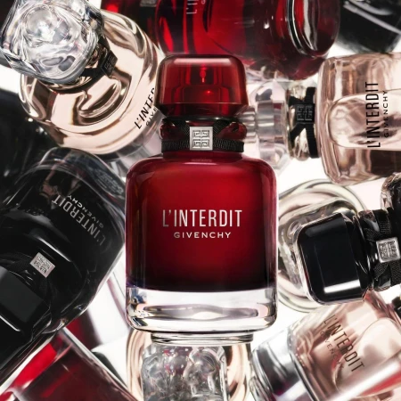Close-up of a red Givenchy L'Interdit perfume bottle surrounded by other assorted fragrance bottles on a white background.