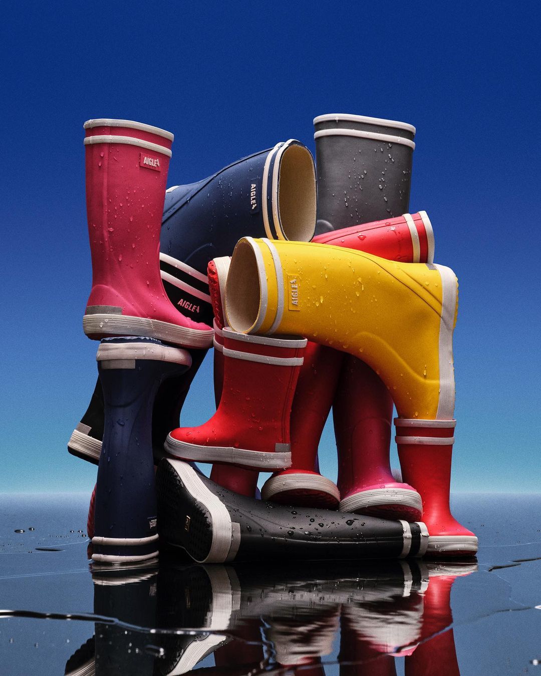 Colorful rubber rain boots arranged in a stacked display against a graduated blue background. Rubber boots in yellow, red, navy, pink and grey.