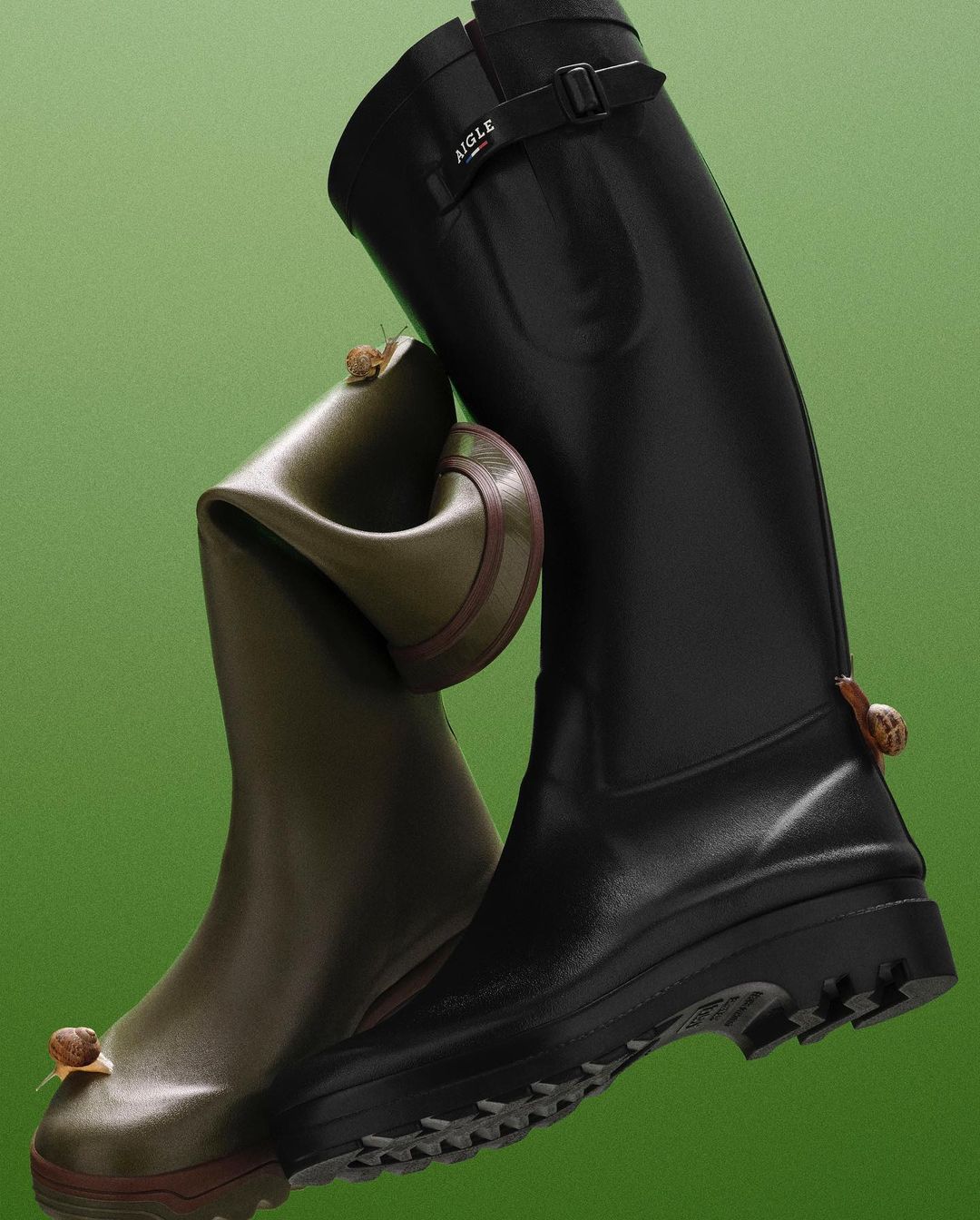 Two rain boots, one black and one olive green, are positioned next to each other with snails crawling on them, set against green background.