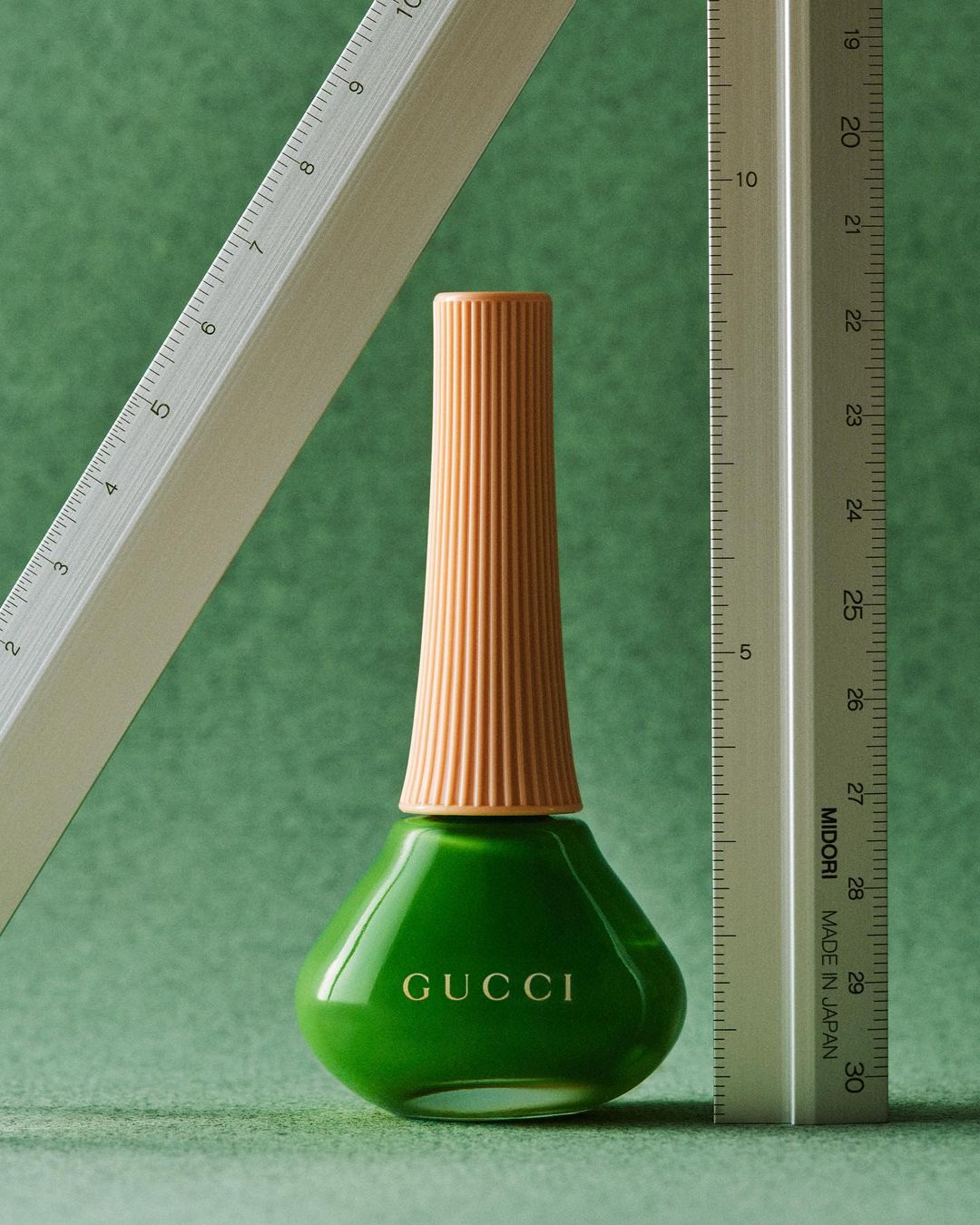 Gucci nail polish bottle in bright green with a ribbed orange neck, flanked by two silver metal rulers on a green background.