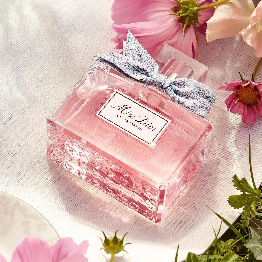 Miss Dior Eau de Parfum bottle on a white cloth surrounded by pink cosmos flowers and green leaves, creating a soft and elegant beauty still life.
