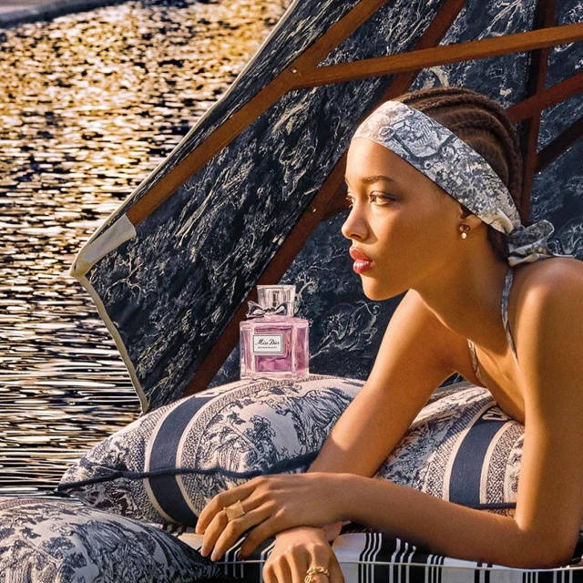 Elegant woman in patterned headband poses beside Miss Dior perfume bottle on a patterned sunbed near water. Beauty and fashion advertisement.