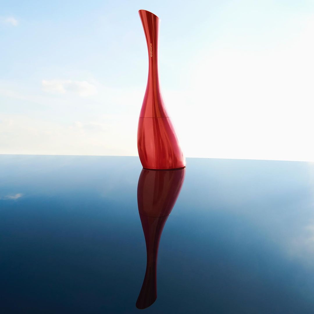Red perfume bottle on a reflective surface against a bright sky background, studio shot, product photography.