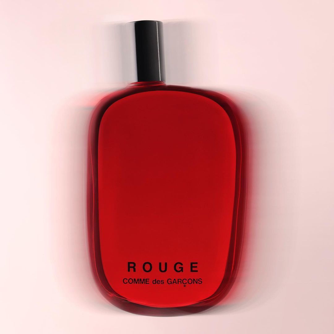 A red glass perfume bottle from Comme des Garçons with a black cap is isolated against a gradient light pink background.