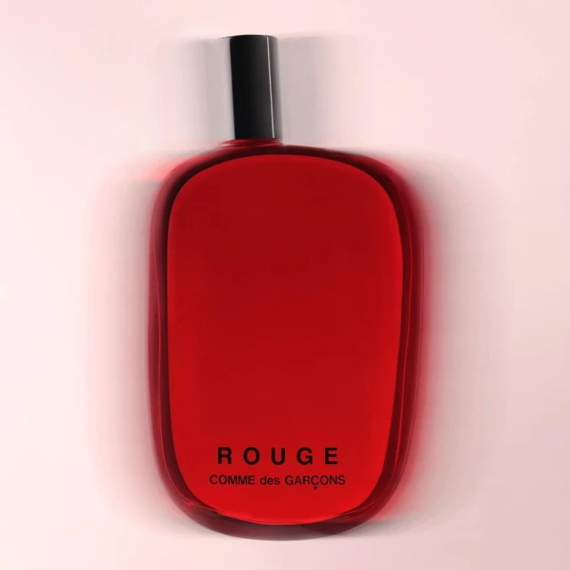 A red glass perfume bottle from Comme des Garçons with a black cap is isolated against a gradient light pink background.