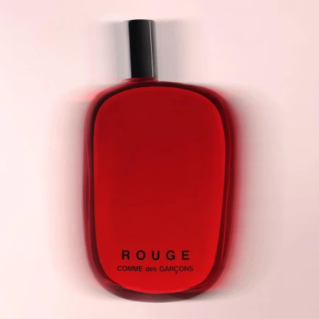 A red glass perfume bottle from Comme des Garçons with a black cap is isolated against a gradient light pink background.