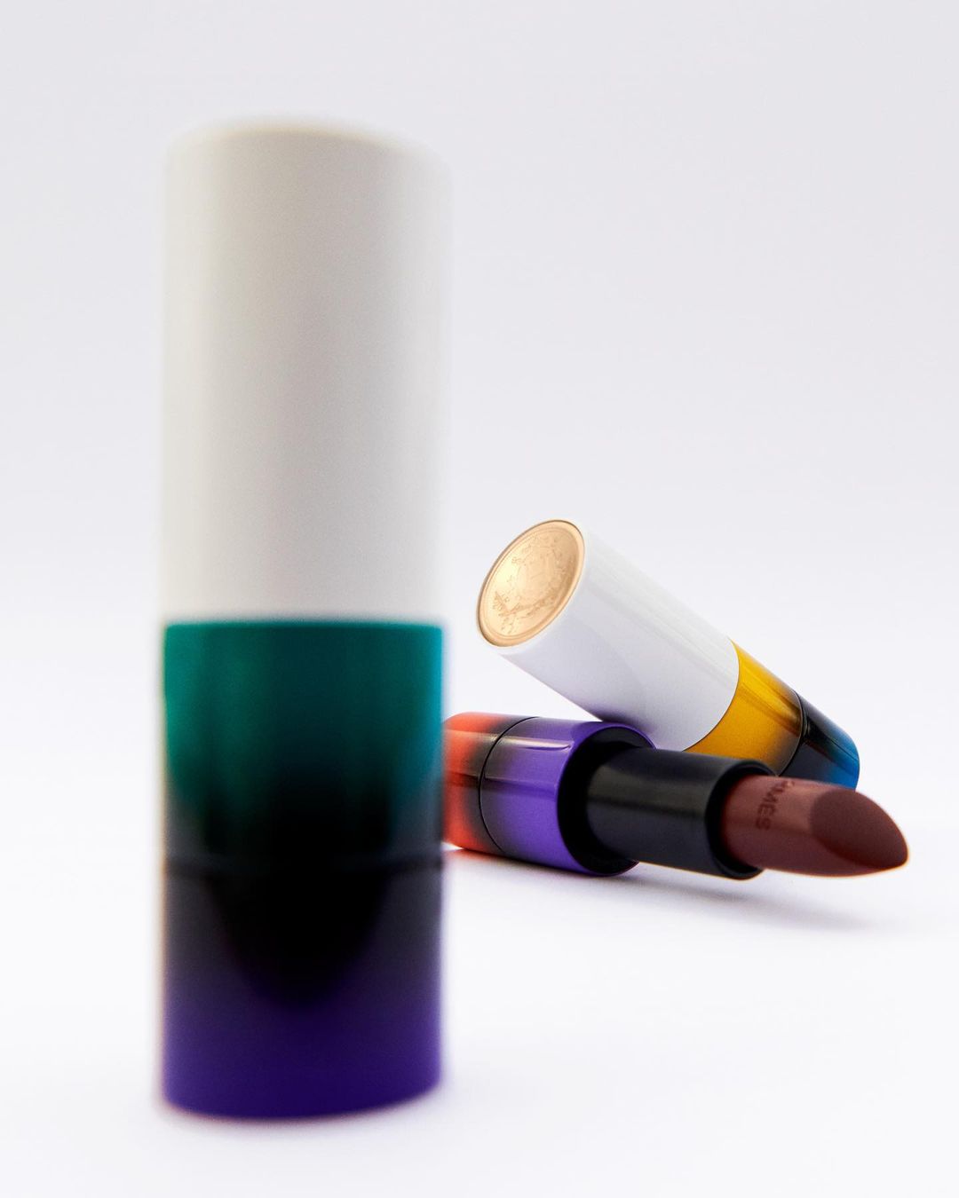 Close-up studio shot of two lipsticks: one standing upright in a white, green, and purple case, and another lying down.