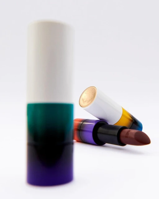 Close-up studio shot of two lipsticks: one standing upright in a white, green, and purple case, and another lying down.