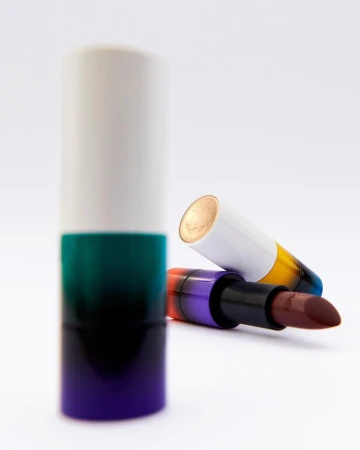 Close-up studio shot of two lipsticks: one standing upright in a white, green, and purple case, and another lying down.