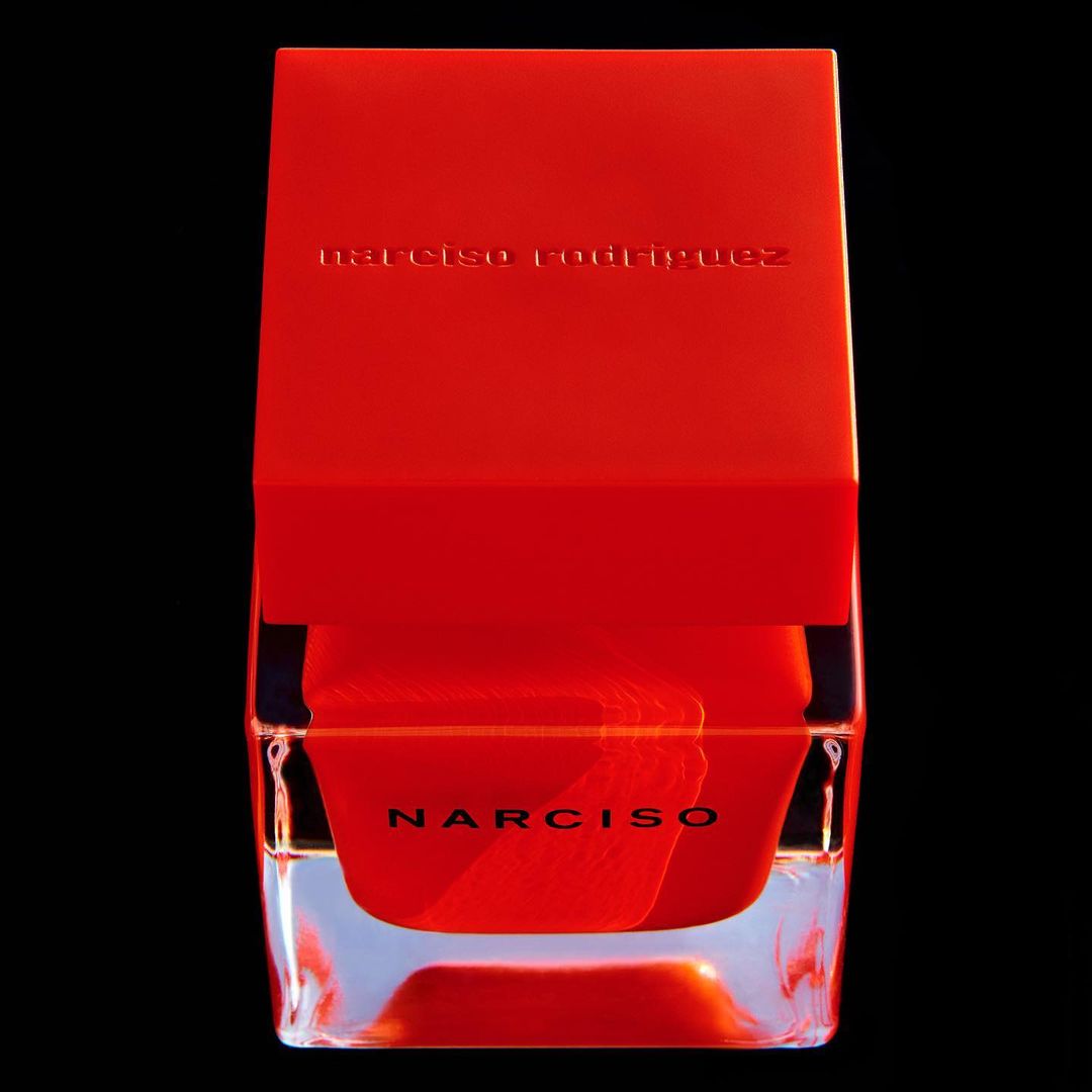 Close-up of a bright red Narciso Rodriguez perfume bottle with a clear base against a dark background in a studio-style product shot.