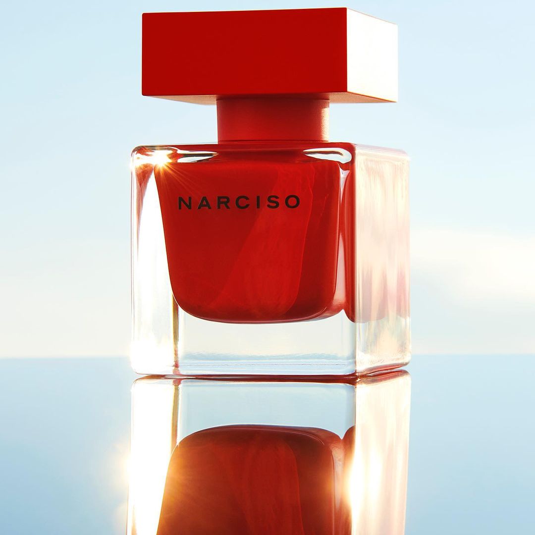 Close-up studio shot of a Narciso Rodriguez red perfume bottle, emphasizing its square design and clear reflections against a blue sky.
