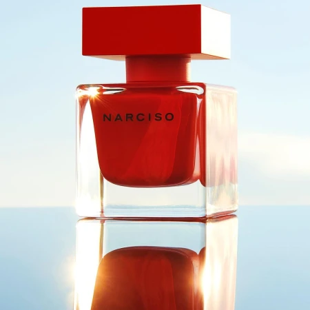 Close-up studio shot of a Narciso Rodriguez red perfume bottle, emphasizing its square design and clear reflections against a blue sky.