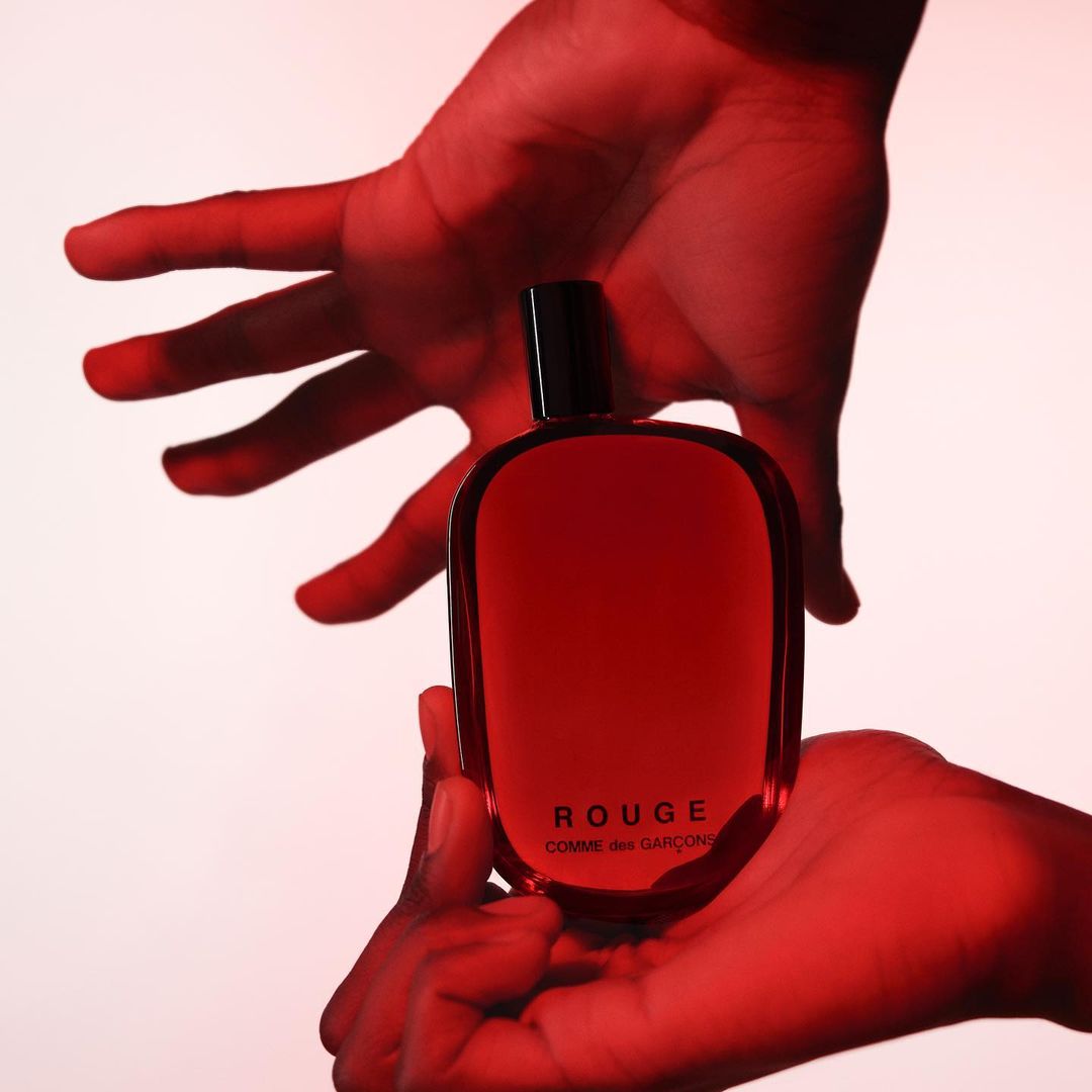 Perfume bottle held in two hands against a soft white backdrop, bathed in a vibrant red light, showcasing its sleek design.