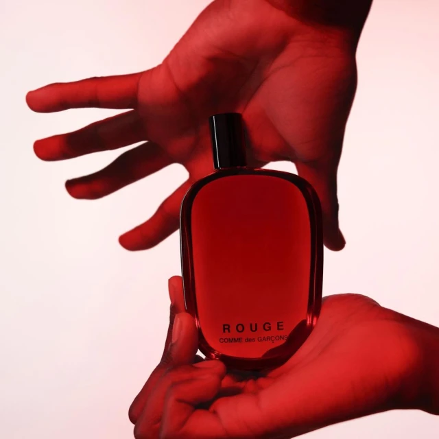 Perfume bottle held in two hands against a soft white backdrop, bathed in a vibrant red light, showcasing its sleek design.