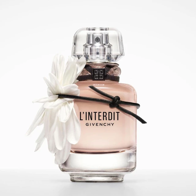 Close-up studio shot of a light pink L'Interdit Givenchy perfume bottle adorned with a white flower and black ribbon on a white backdrop.