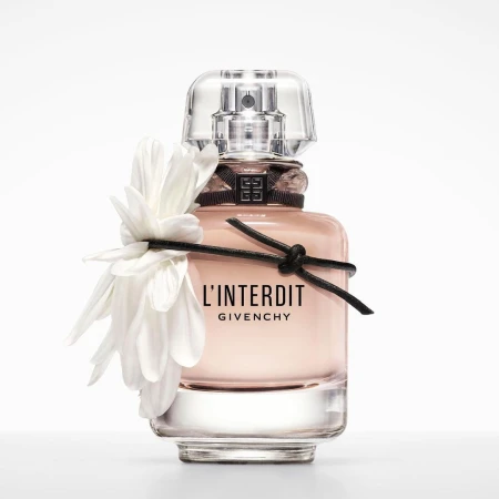 Close-up studio shot of a light pink L'Interdit Givenchy perfume bottle adorned with a white flower and black ribbon on a white backdrop.
