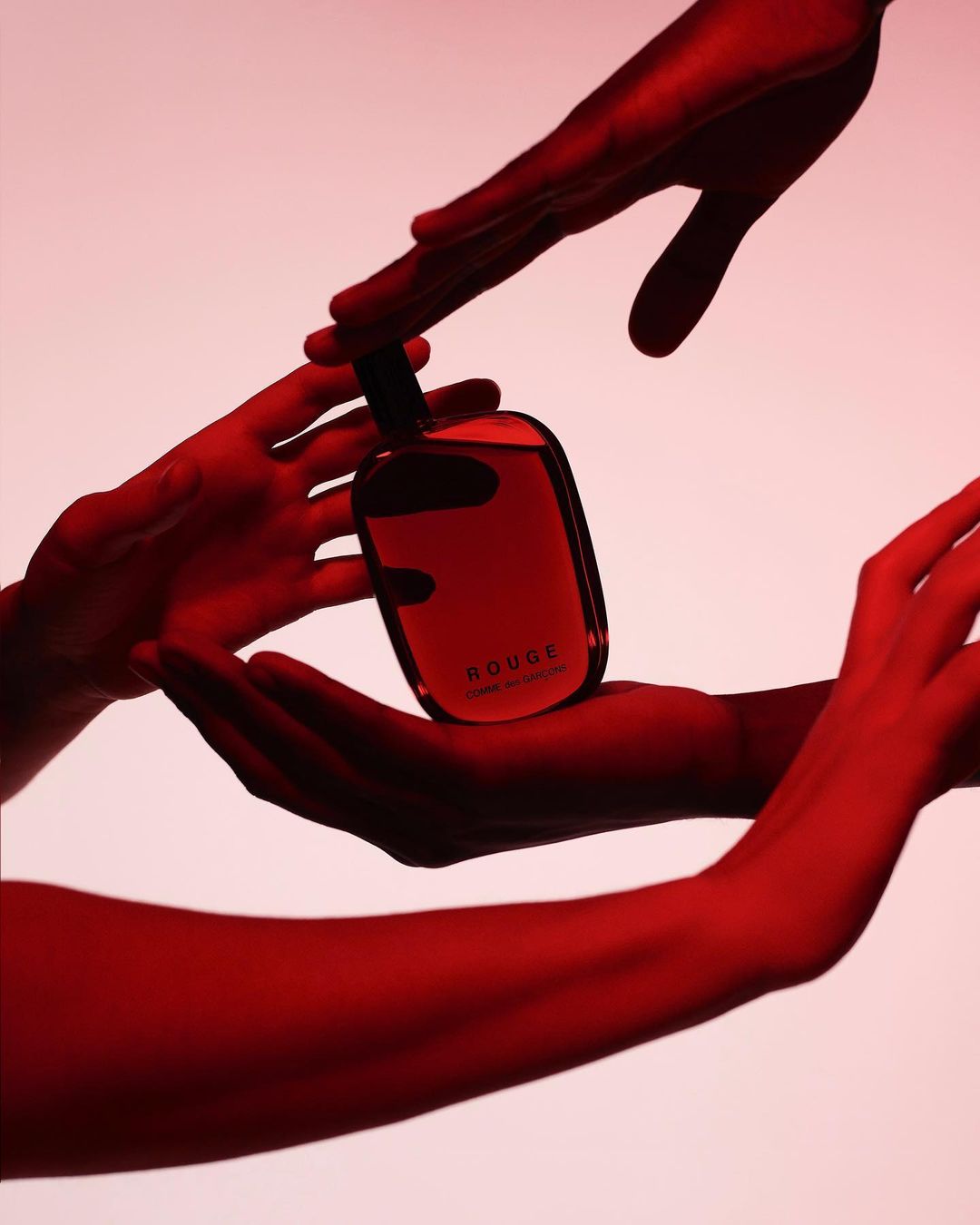 Close-up of a glass perfume bottle with a black cap being held by multiple hands in a studio setting with red lighting.