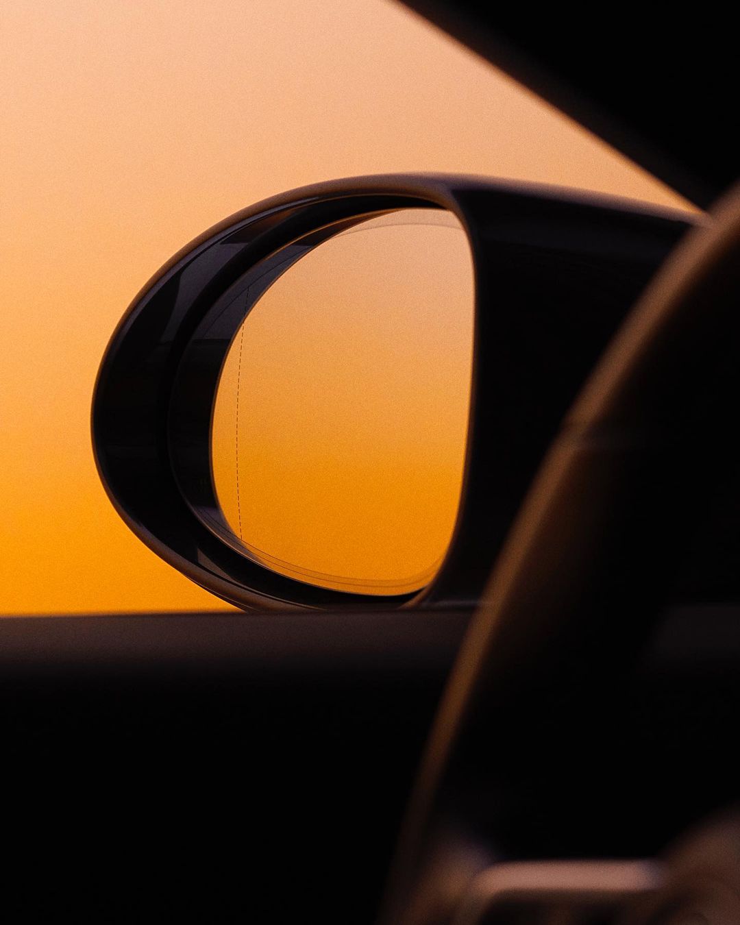 Car side mirror reflecting orange sunset sky, view from inside vehicle. Automotive interior detail with sky reflection.