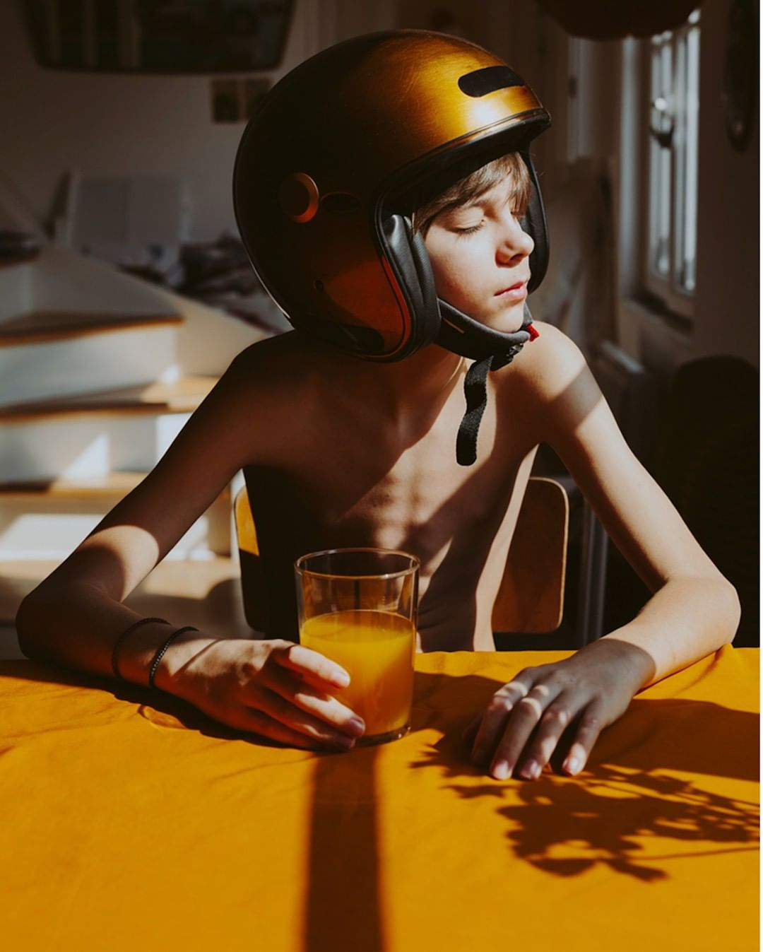 Boy with helmet sitting at a table with orange juice, bathed in natural light, creating a youthful, playful yet contemplative mood.