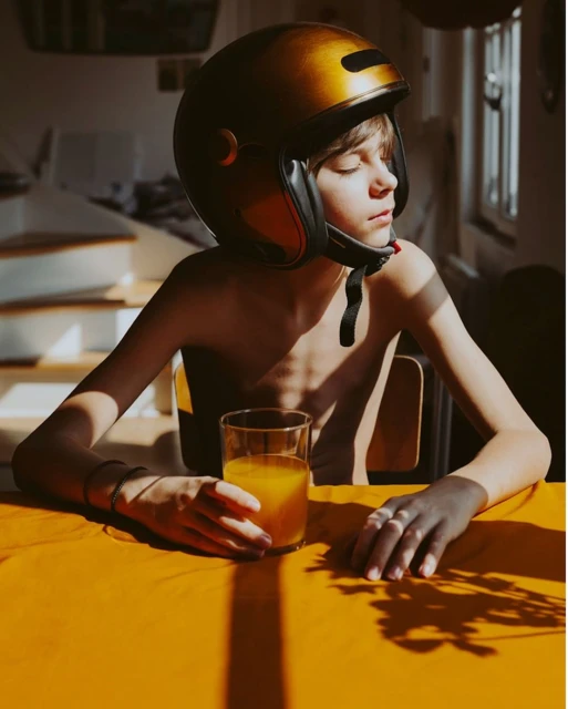 Boy with helmet sitting at a table with orange juice, bathed in natural light, creating a youthful, playful yet contemplative mood.