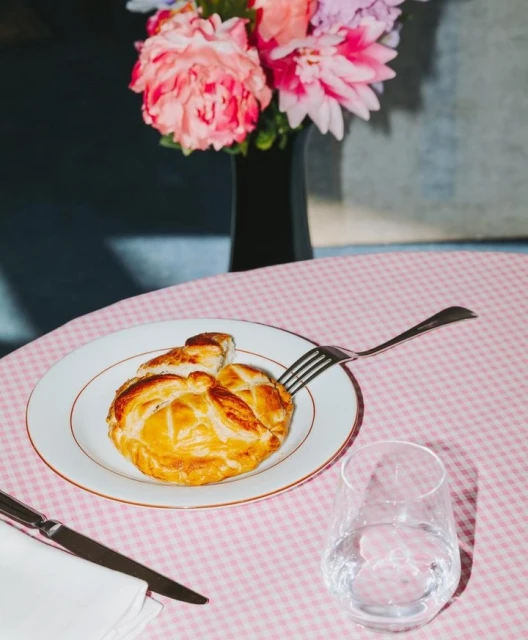 Golden baked pastry on a white plate with a fork and knife on a pink gingham tablecloth, featuring a vase of flowers.