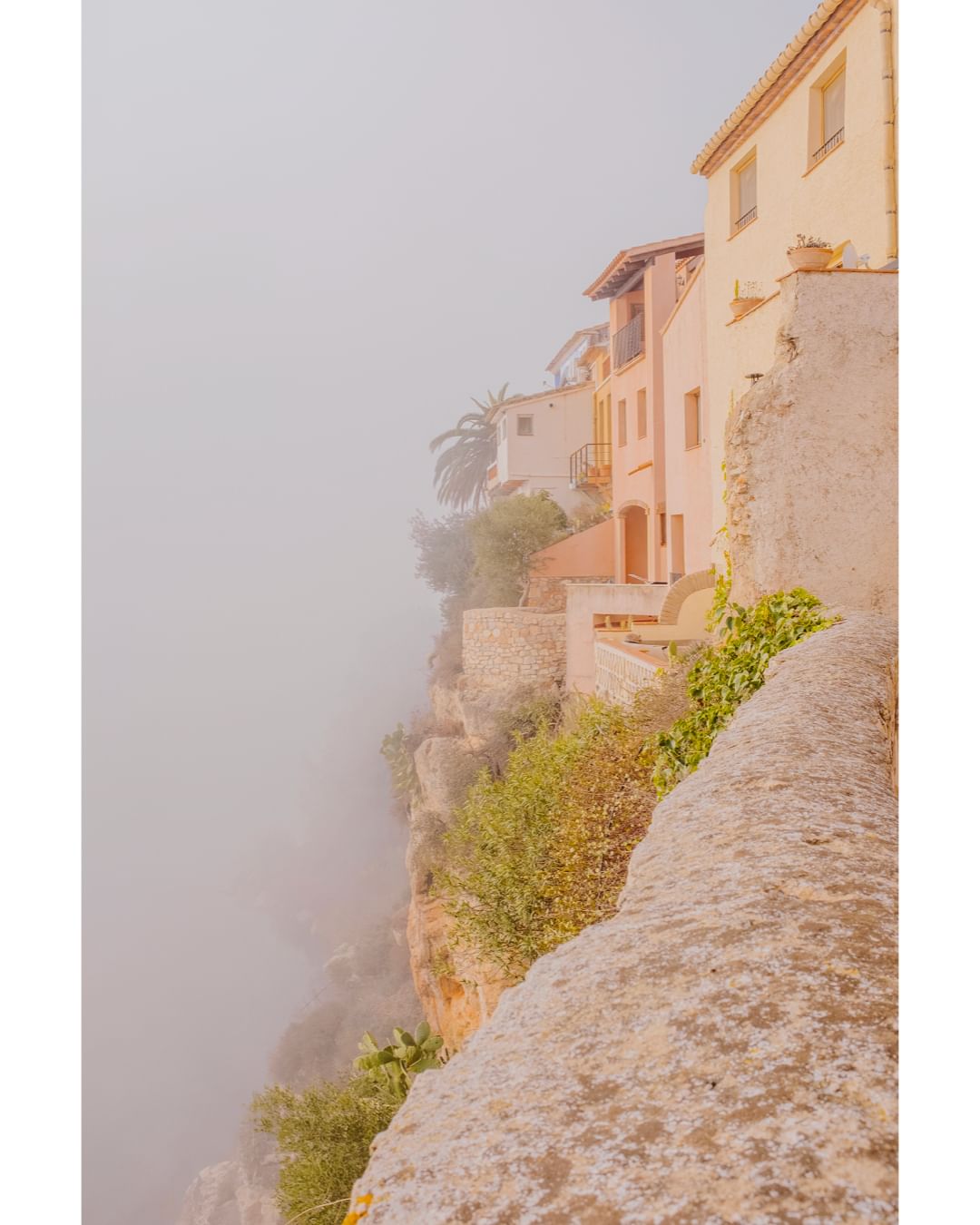 Picturesque view of stone buildings on a cliffside shrouded in mist, featuring lush greenery and soft, warm tones. Travel destination.
