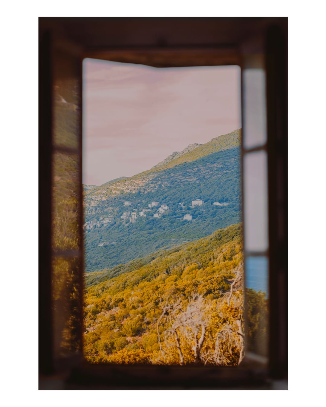 A view of a mountainside filled with trees seen through an open wooden window, creating a natural, framed vista.