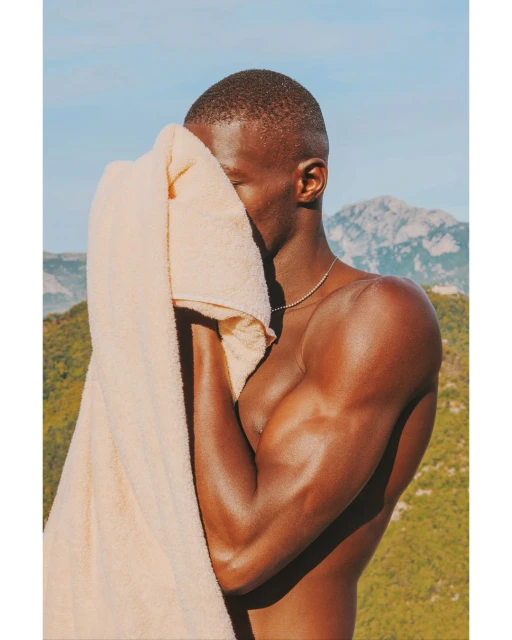 Shirtless man with tan complexion holds a towel to face in an eye-level shot in front of a mountain and blue sky.